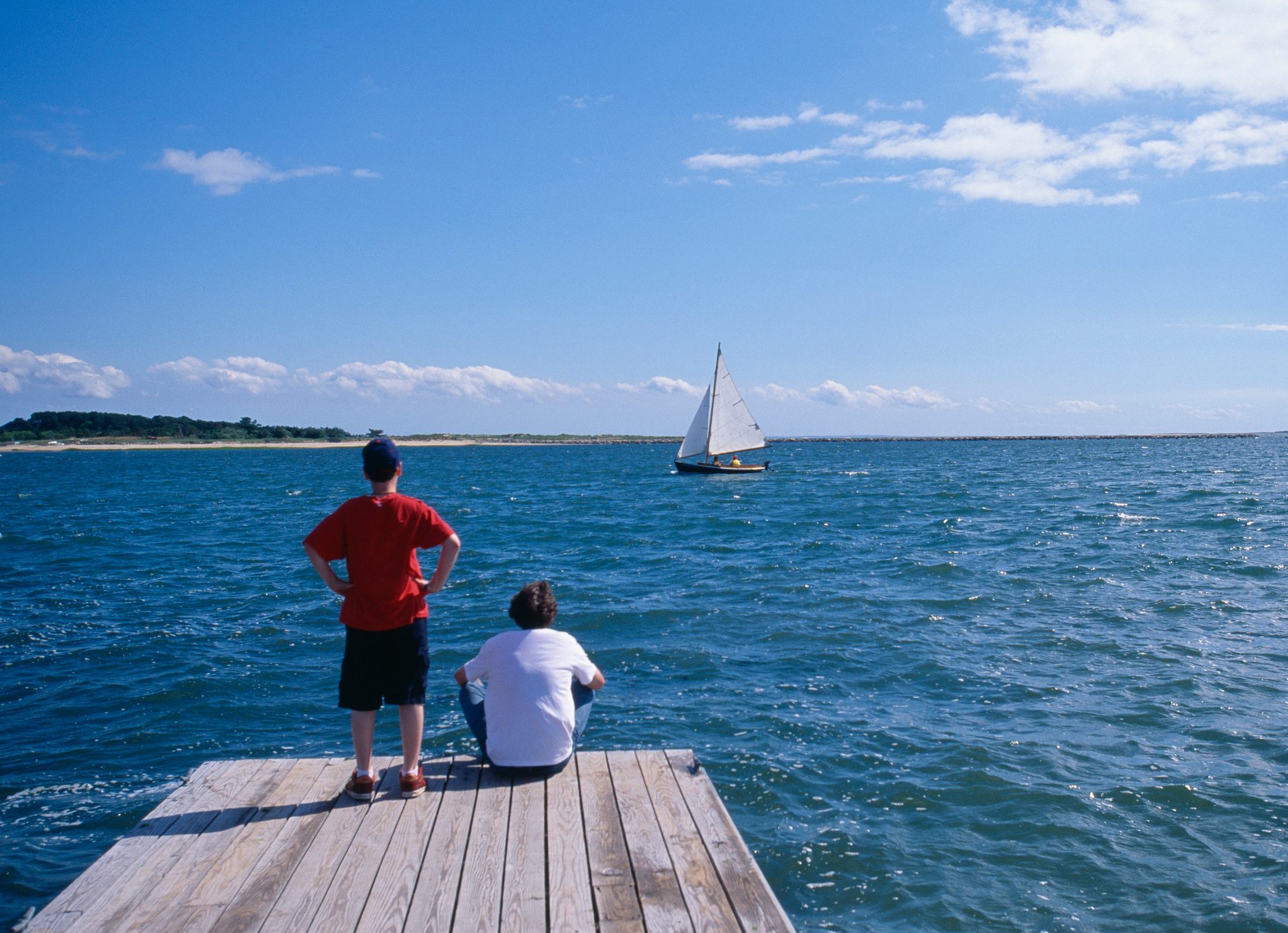Two boys sitting on dock, watching sailboat, rear view