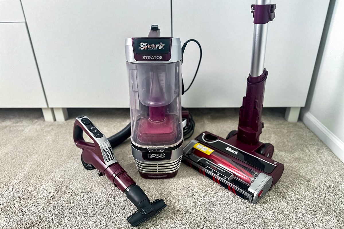 A disassembled vacuum cleaner with parts spread out on a carpeted floor. The components include the main unit, a handle with a hose, and a detachable floor brush. The vacuum is predominantly maroon and black.