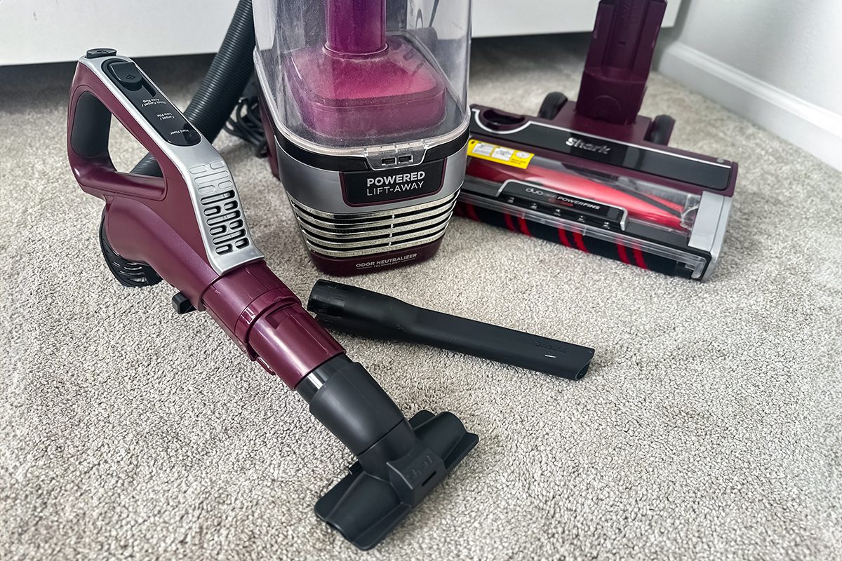 A red and gray vacuum cleaner with detachable parts is displayed on a light carpet. The vacuum features a canister, a hose with a handle, and various attachments, including a narrow nozzle and a brush head.
