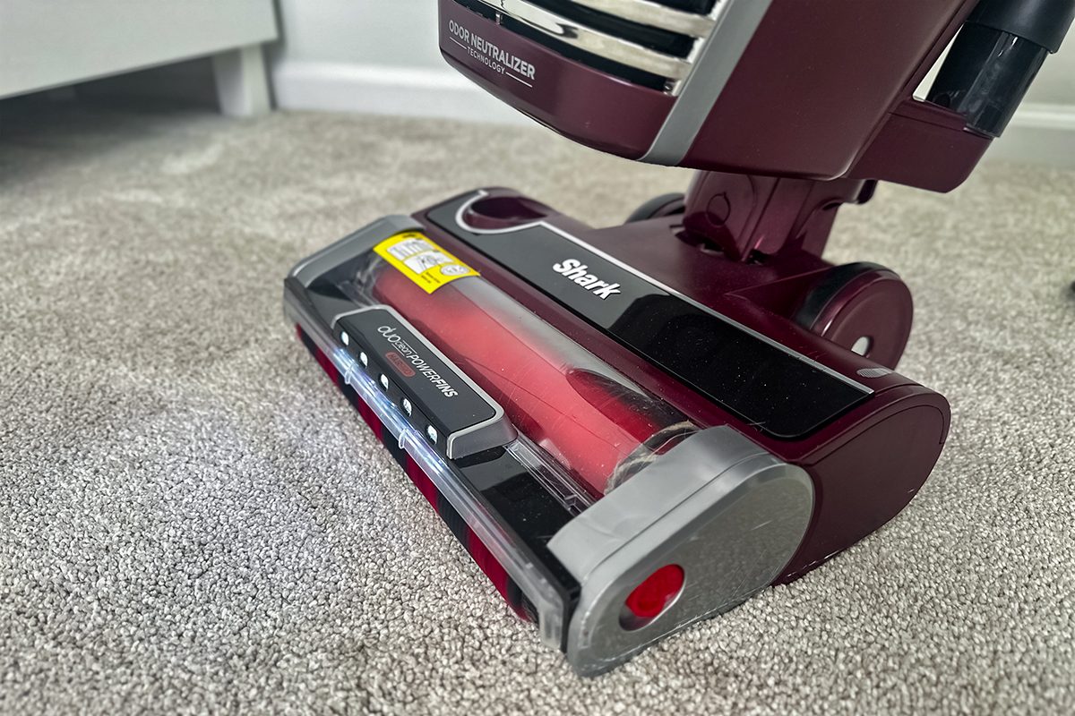A maroon vacuum cleaner on a gray carpet, displaying the brush roll component. The vacuum cleaner is a Shark brand, with a sleek design and clear protective cover over the brush.