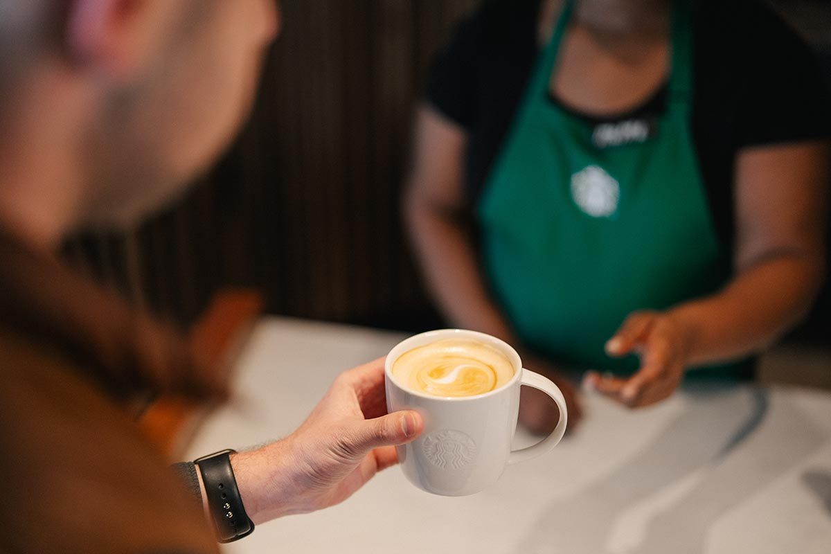 Starbucks barista in green apron hands a coffee drink in a ceramic mug to a customer