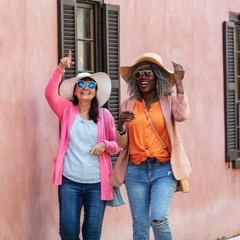 Two women in sunglasses and sun hats walk in front of a dark orange building