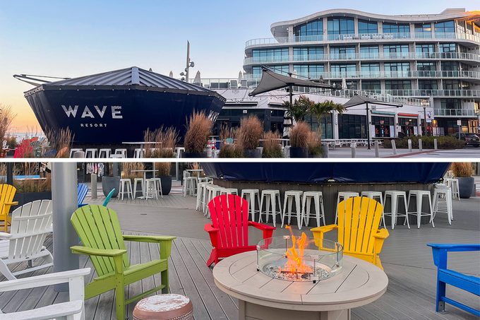 Outdoor seating area with colorful Adirondack chairs surrounding a fire pit. Behind, a modern building with balconies and a large structure labeled "WAVE" on the facade. Tables and stools are arranged around the space.