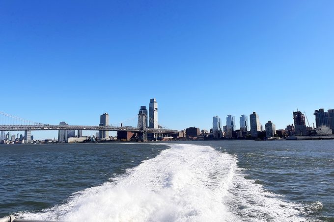 View from a boat leaving a frothy wake in the water, with a city skyline under a clear blue sky in the background. A bridge spans the river, connecting to tall modern buildings.