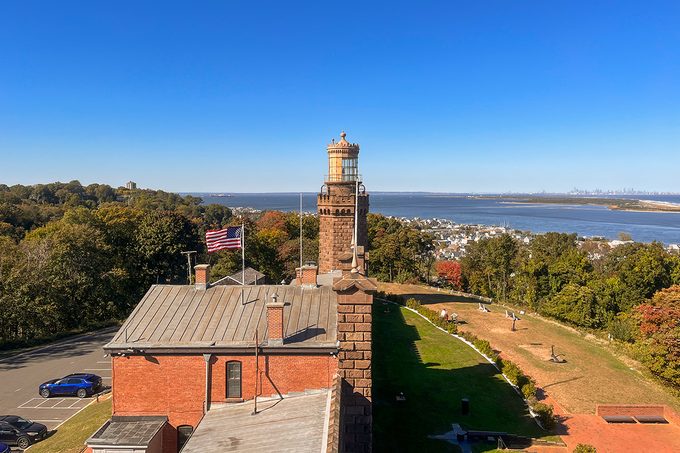 The image shows a historic brick lighthouse with a large building in the foreground, surrounded by trees. An American flag waves in the breeze. In the background, there is a view of a river and distant land under a clear blue sky.