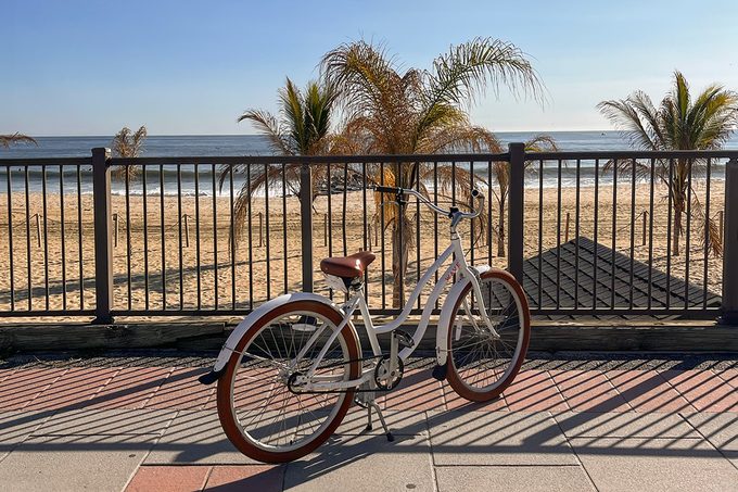 A white bicycle with brown accents is parked on a paved walkway by a metal railing. Behind the railing, there is a sandy beach with several palm trees and a calm ocean under a clear blue sky.
