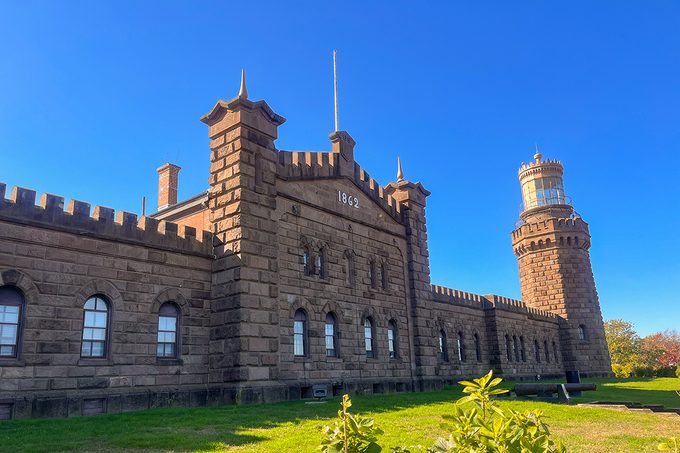 A historic stone building with crenellated roof and a tall lighthouse tower on the right, set against a clear blue sky. The structure is surrounded by grass and a few scattered plants.
