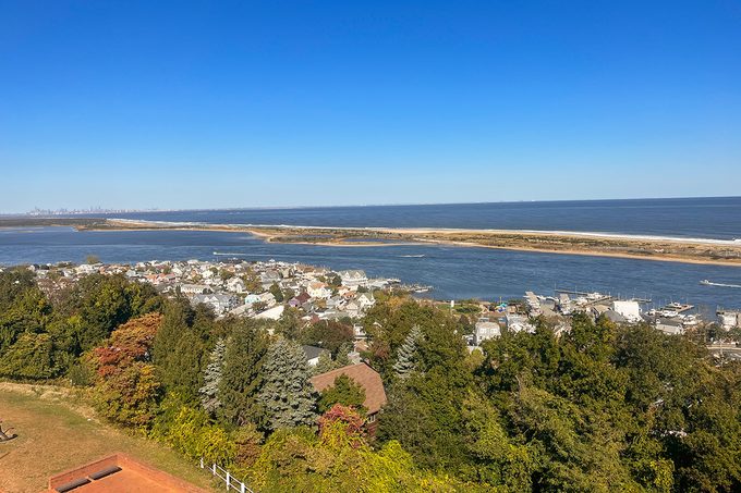 Aerial view of a coastal town surrounded by dense green foliage. The town sits adjacent to a vast body of water, with a long, sandy stretch separating the water from the open sea. Clear blue sky above.