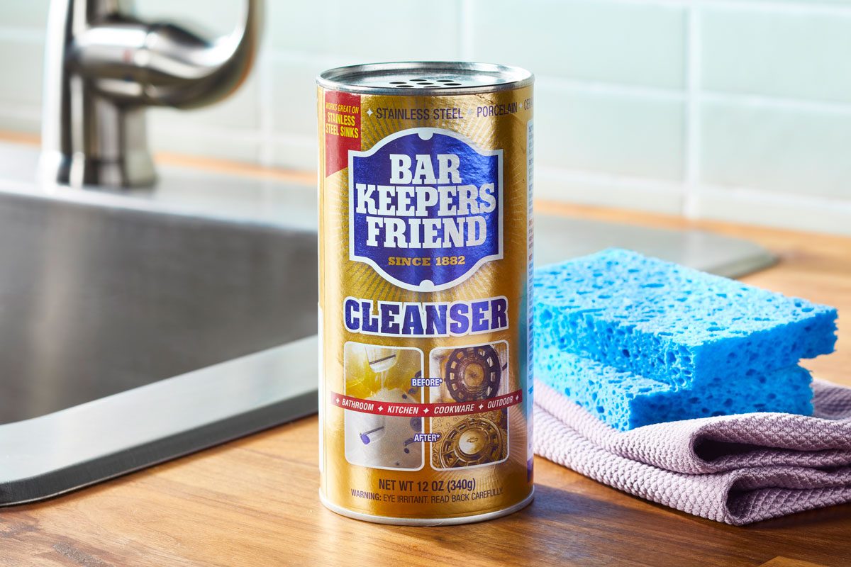 bottle of bar keeper's friend on a kitchen counter near sink, next to sponges and cleaning cloth