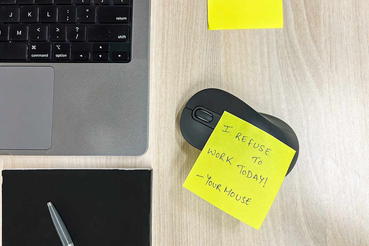 A laptop keyboard is partially visible on a wooden desk. A sticky note on a mouse reads, "I refuse to work today! - Your Mouse." A pen lies on a closed notebook, with another sticky note above them.