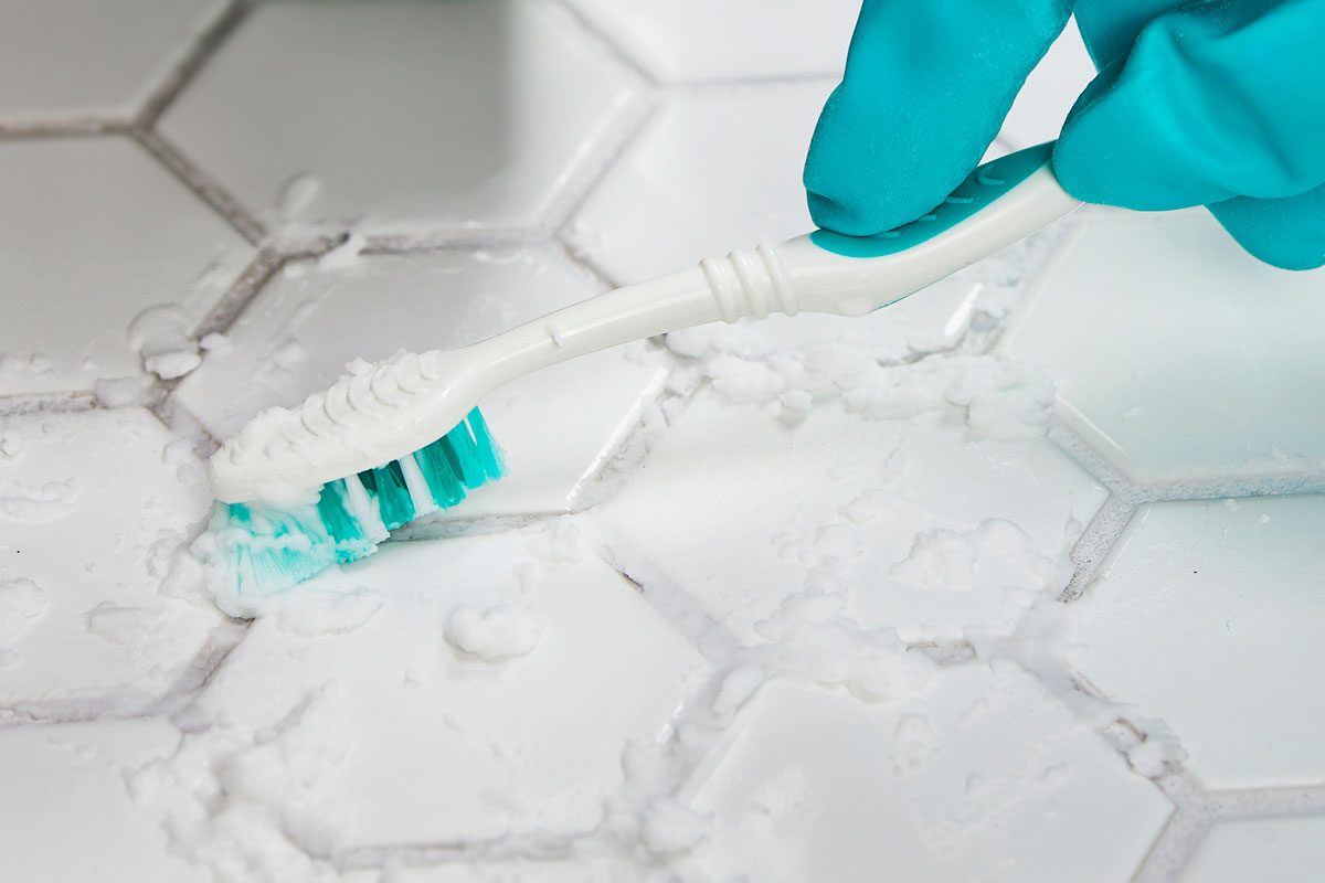 Tight crop of gloved hand using a toothbrush to scrub grout on a white tiled floor