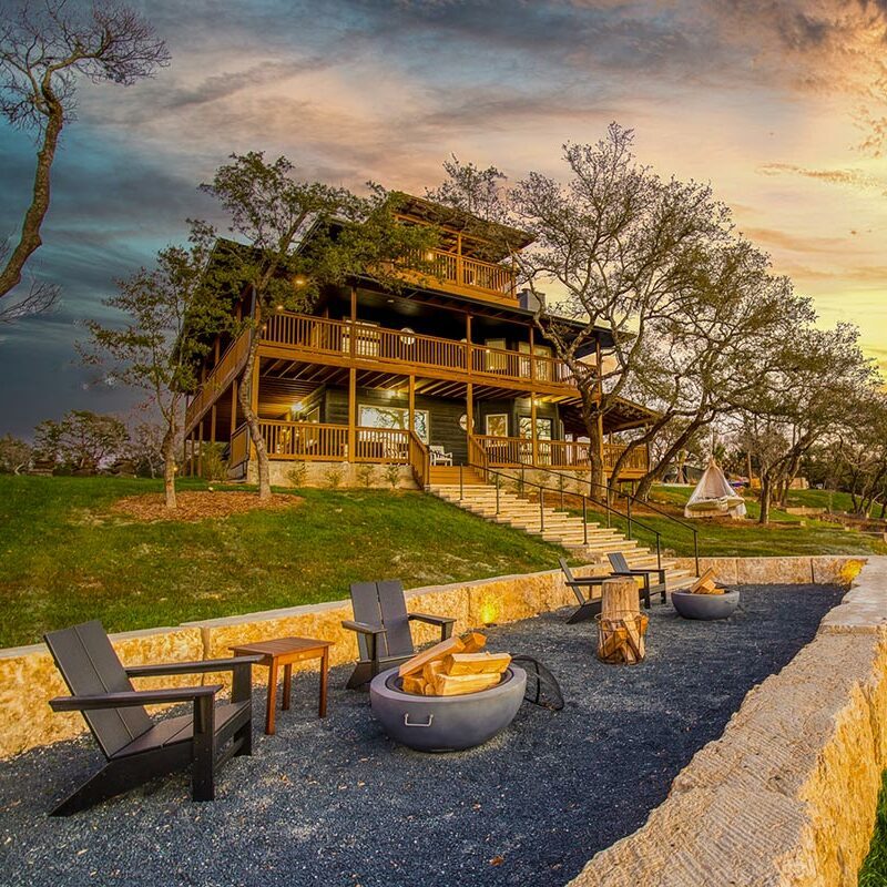Three-story cabin-themed house with a fire pit and adirondack chairs on a hillside at sunset