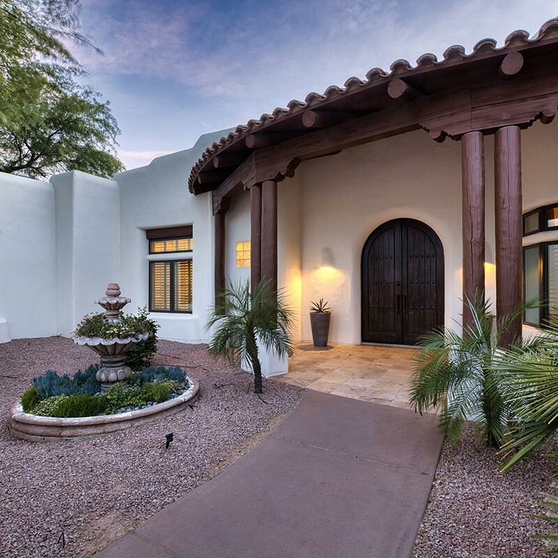 Entrance to a white, Adobe-style home with a wooden awning and a fountain in the front yard